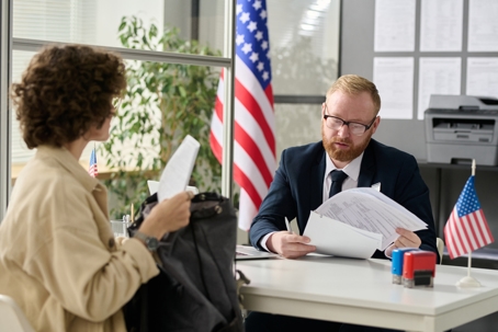 USCIS officer reviewing documents with an applicant during an immigration interview at an office with American flags visible. | Monument Immigration